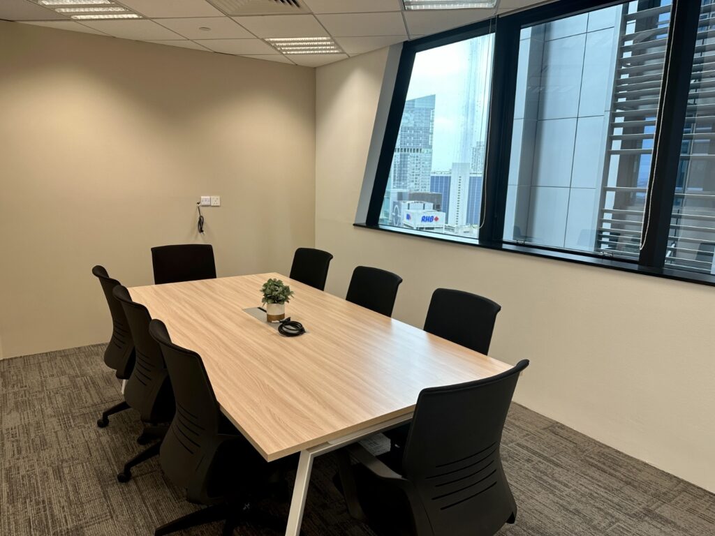 A modern conference room with an empty rectangular table, eight black chairs, a small plant centrepiece, and large windows showing city buildings outside.