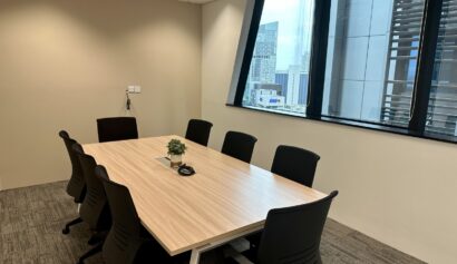 A modern conference room with an empty rectangular table, eight black chairs, a small plant centrepiece, and large windows showing city buildings outside.
