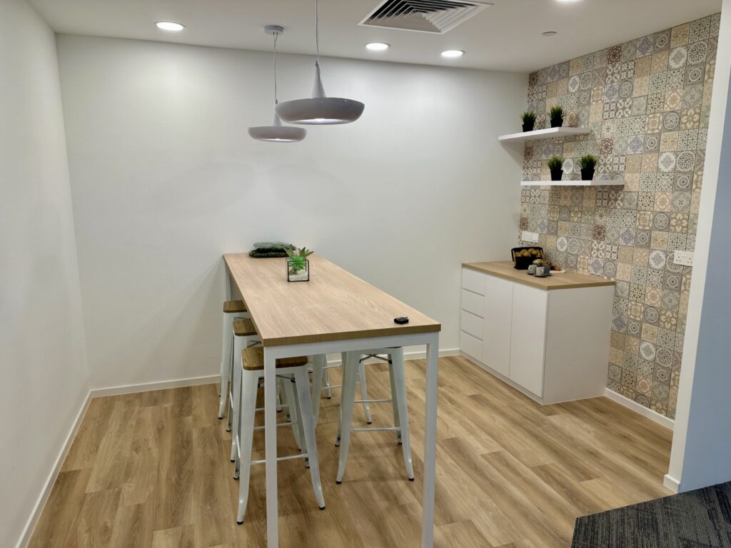 A modern kitchenette with a wooden table, four stools, white cabinetry, potted plants, pendant lights, and a wall decorated with patterned tiles.