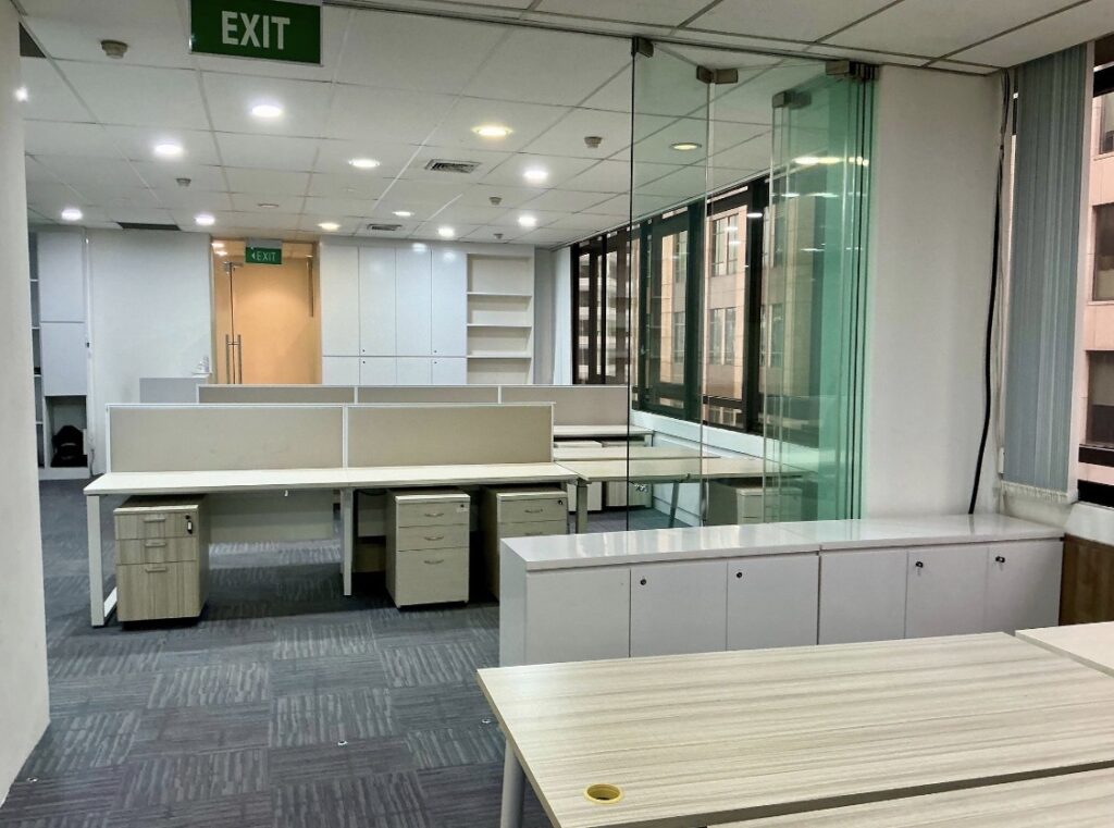 Modern office space with empty desks, storage cupboards, glass partition, large windows, and ceiling lights; an exit sign is visible above the doorway.