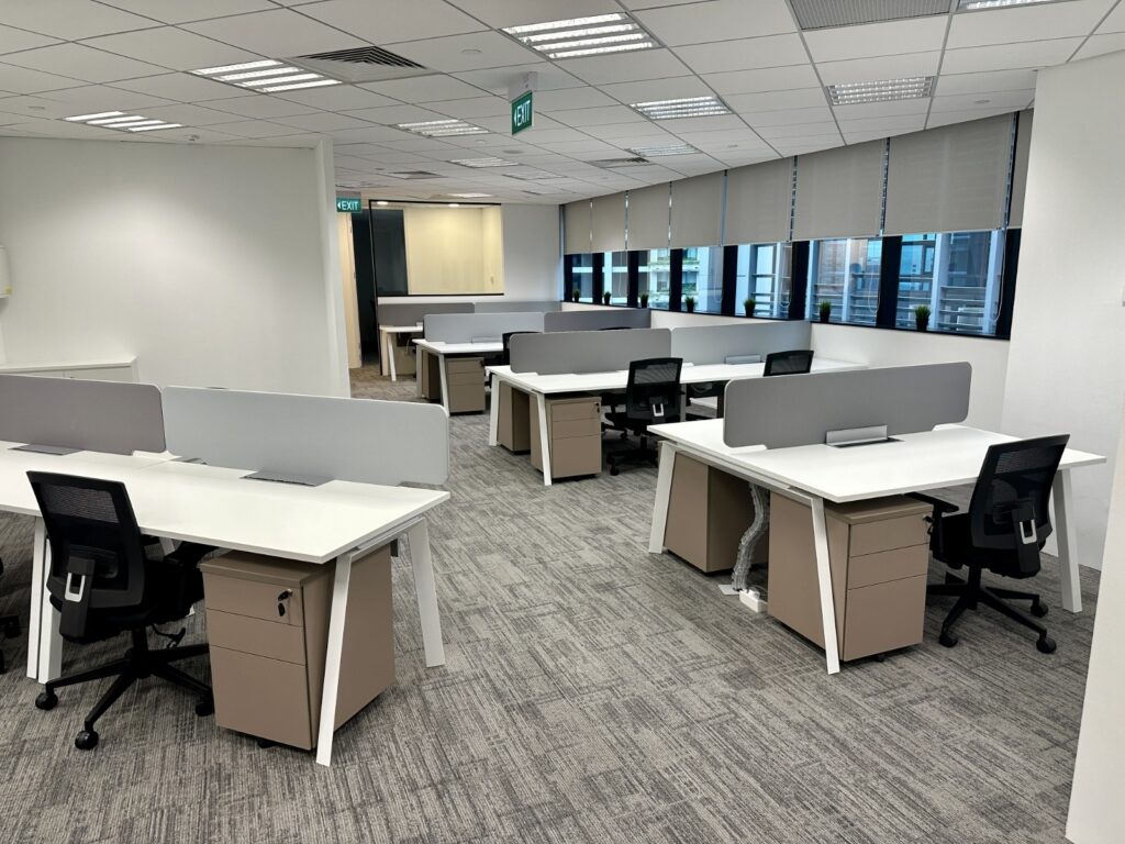 Open office space with several empty desks and chairs, grey partitions, carpeted floor, and large windows with blinds partially lowered.