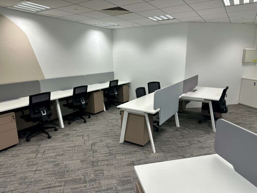 Modern office space with several empty desks, partition screens, office chairs, and neutral-coloured walls and carpet.