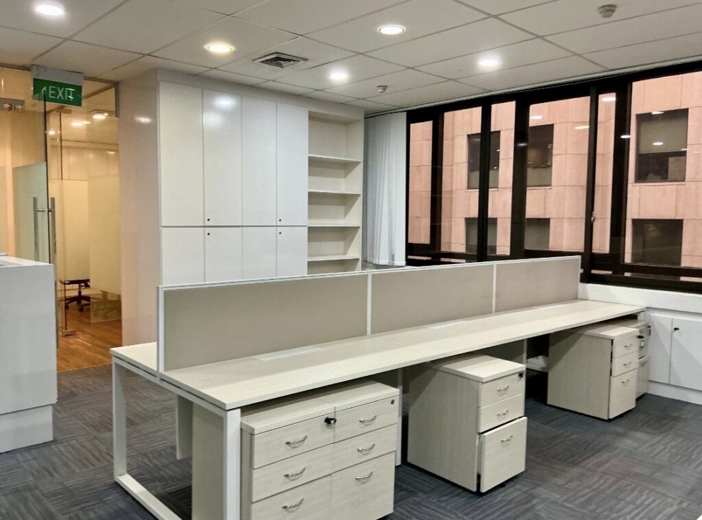 Modern office space with white desks, cupboards, drawers, and a partition, featuring large windows and overhead lighting. An emergency exit sign is visible in the background.