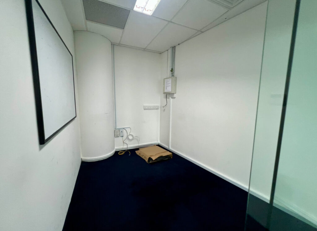 Small empty office with a blank whiteboard, glass wall, carpeted flooring, exposed cables, and a piece of cardboard on the floor near electrical panels.