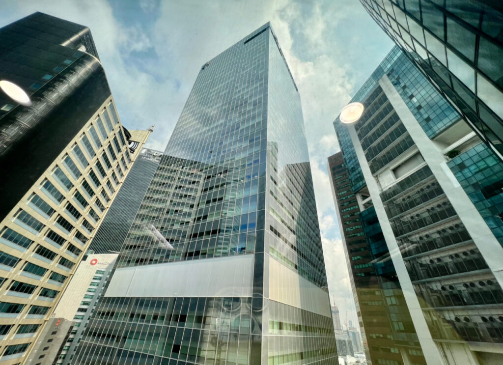 Tall glass office buildings under a partly cloudy sky, viewed from a high vantage point with reflections visible in the window.