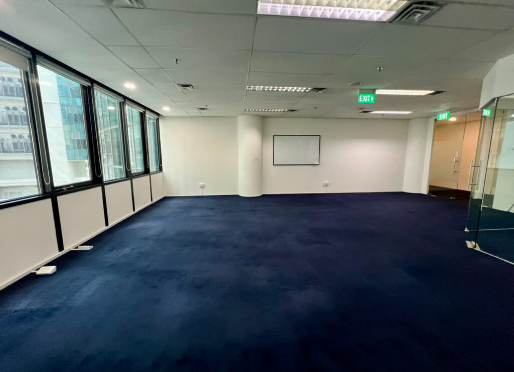 Empty office space with blue carpet, large windows along one wall, a whiteboard on another wall, and glass doors near the exit signs.