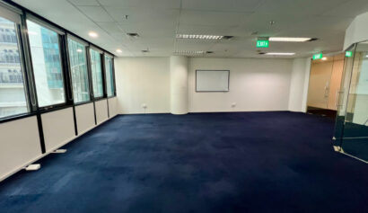Empty office space with blue carpet, large windows along one wall, a whiteboard on another wall, and glass doors near the exit signs.