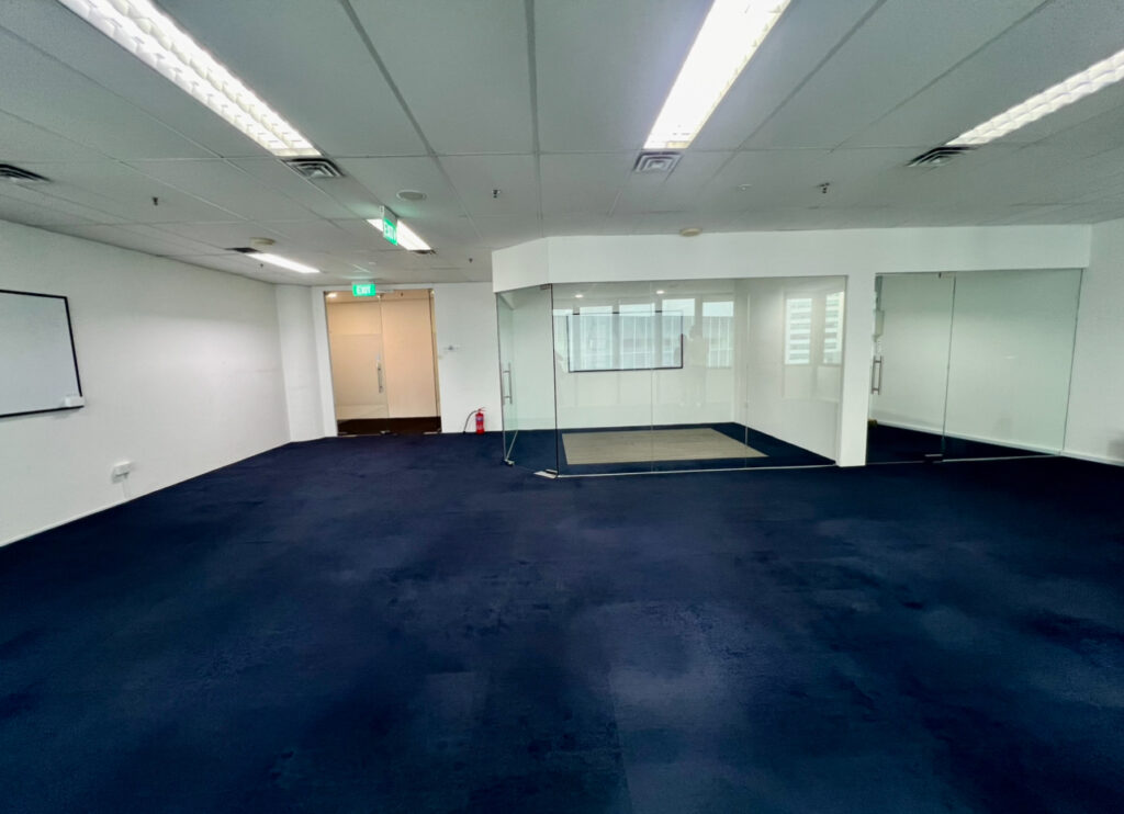 Empty office space with blue carpet, white walls, a whiteboard, glass-walled meeting room, and fluorescent ceiling lights.