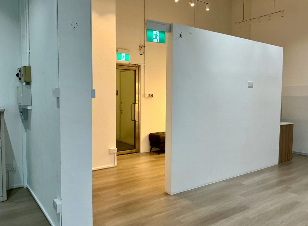 An empty room with white partition walls, wood flooring, and a glass door at the back beneath a green emergency exit sign.