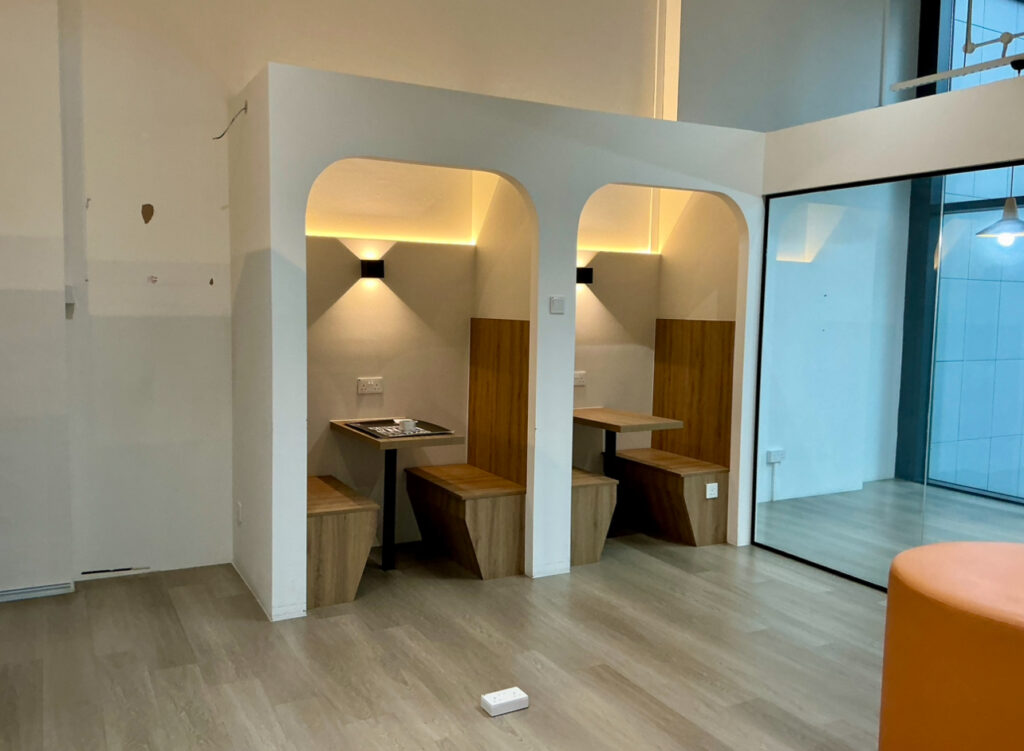 Two small wooden booths with built-in seating and tables are set into a white alcove, illuminated by wall lights. A glass wall and orange seat are visible on the right.