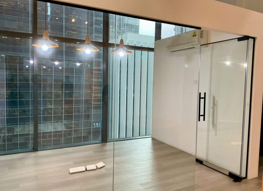 Glass-walled office room with wood flooring, ceiling lights, and air conditioning unit. Large windows show city buildings outside. Several power sockets are on the floor.