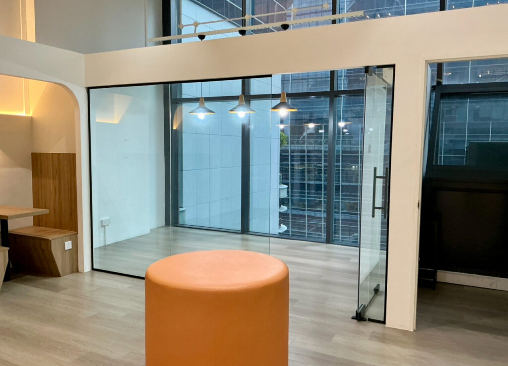 Modern office space with glass-walled meeting room, overhead pendant lights, and an orange cylindrical pouffe in the foreground. Large windows show buildings outside.