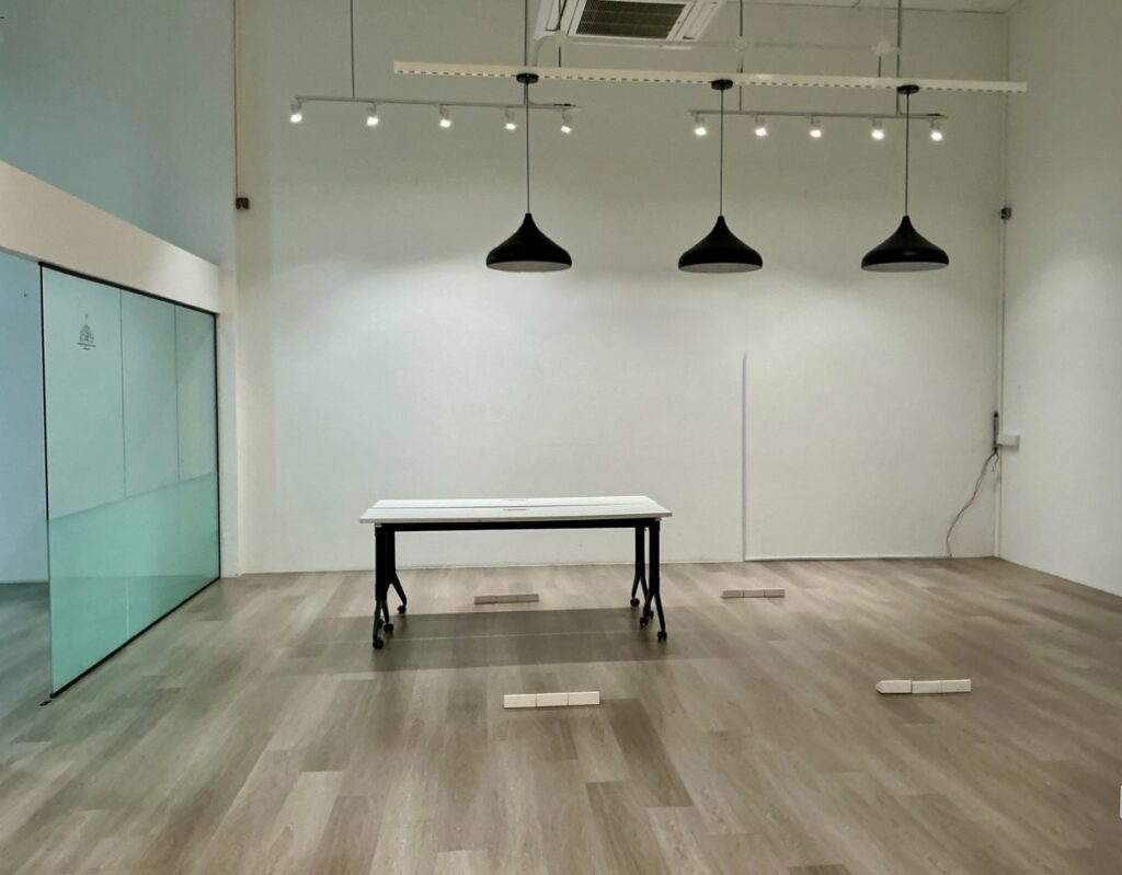 Minimalist room with wood flooring, a single table at the centre, three black pendant lights overhead, white walls, and a large glass panel on the left side.