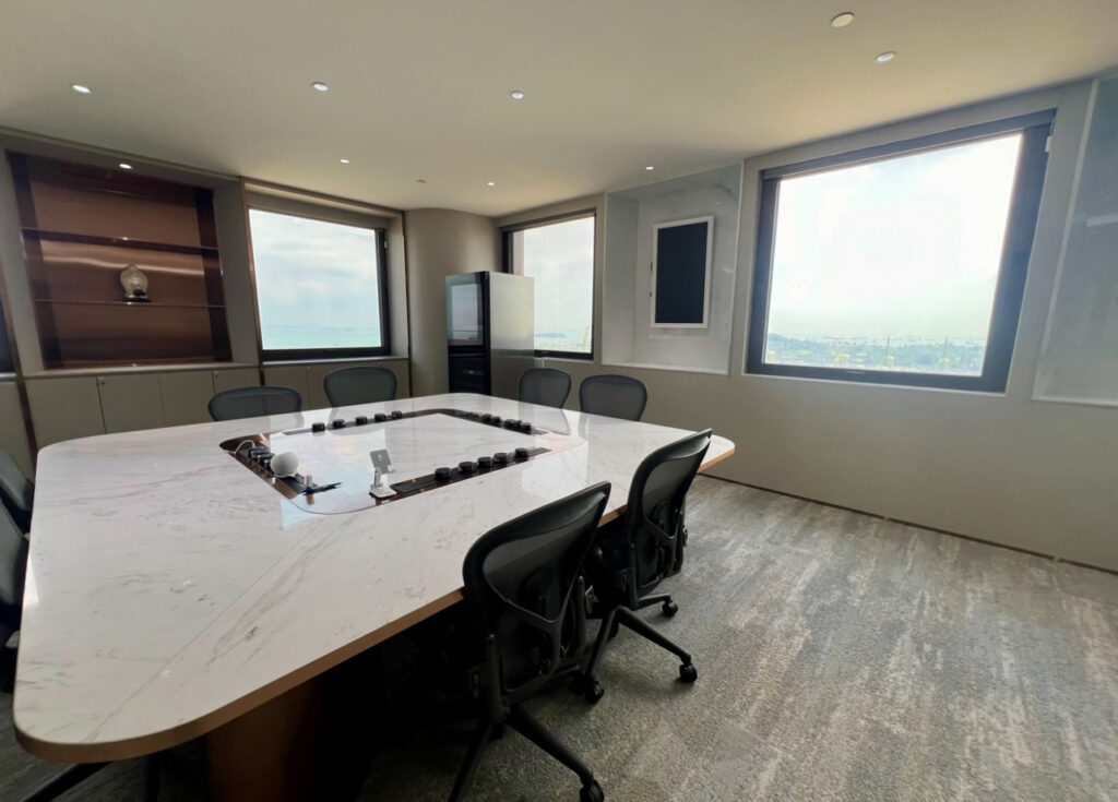 Modern conference room with a large marble table, black chairs, and large windows offering a city view. Shelves and a screen are mounted on the walls.