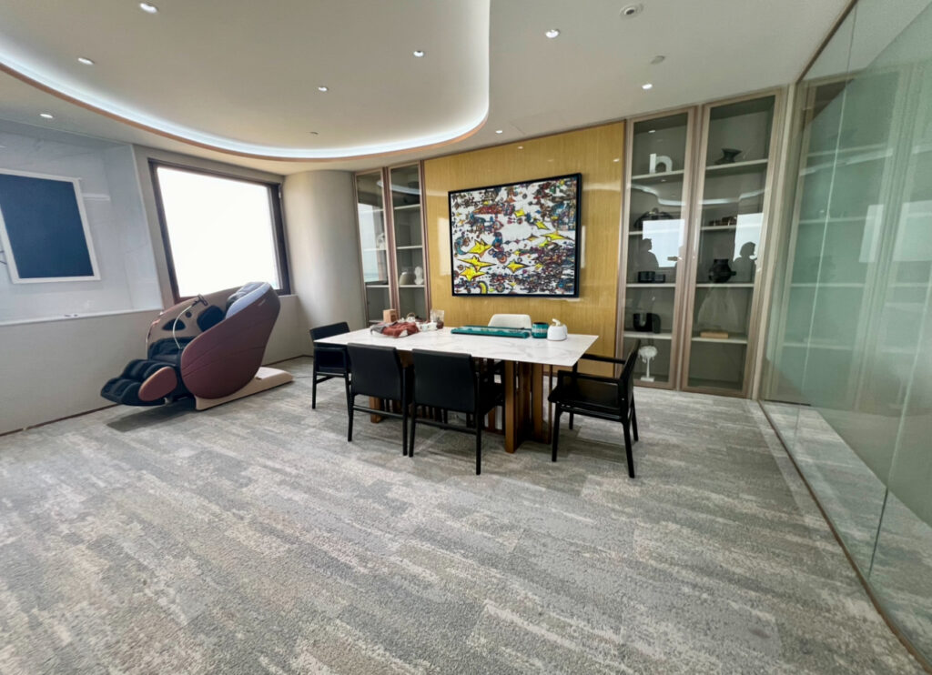 Modern conference room with a rectangular table, six chairs, abstract wall art, glass cupboards, and a brown massage chair in the corner.
