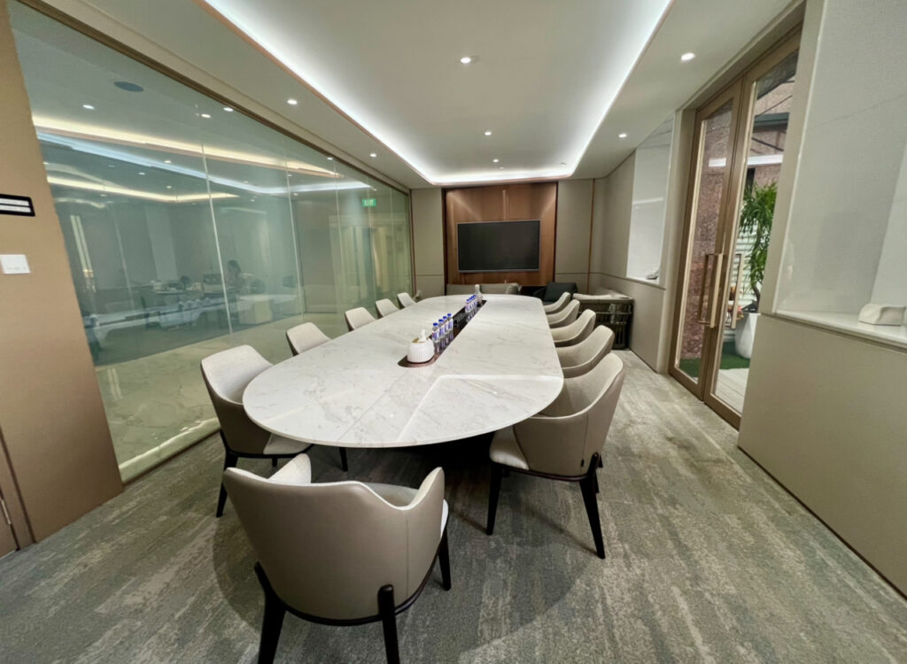 Modern conference room with a long marble table, ten beige chairs, a wall-mounted TV, bottled water, and glass walls.