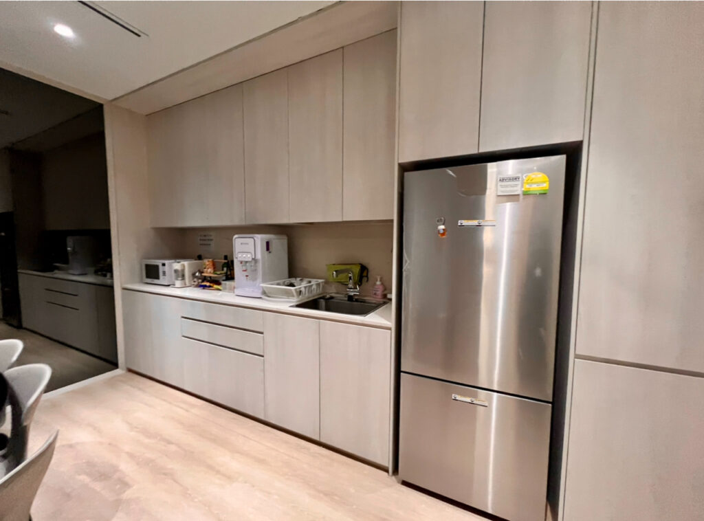 Modern kitchen with light wood cupboards, a stainless steel fridge, microwave, water dispenser, sink, and countertop appliances.