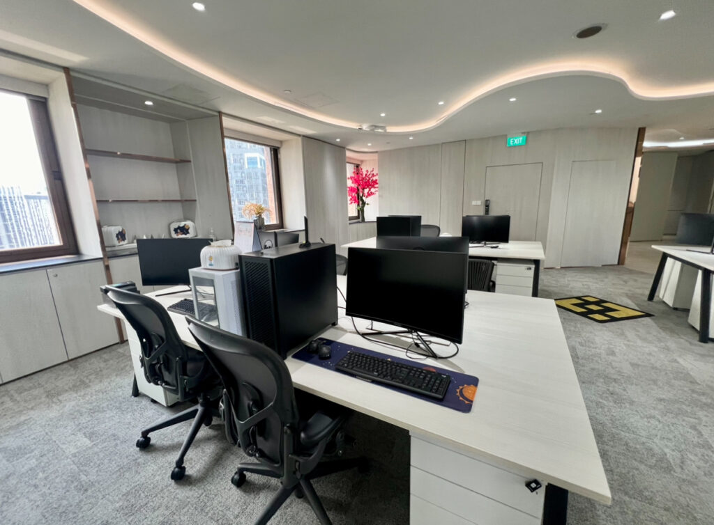 Modern open-plan office with several workstations, ergonomic chairs, desktop computers, and large windows with city views. Bright lighting and neutral decor create a clean, professional space.