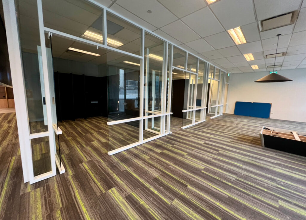 An empty office space with floor-to-ceiling glass-walled partitions, brown and green striped carpet, a suspended pendant light, and a small blue acoustic panel against a white wall.