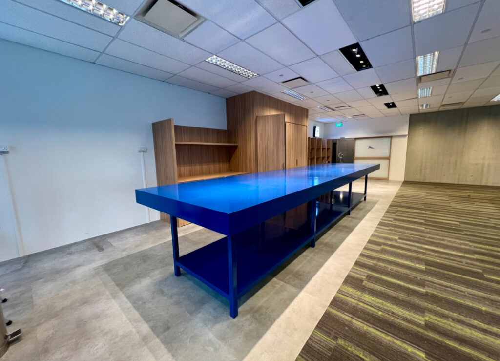 A long blue table with shelves underneath stands in a modern room with wooden cupboards, white walls, and a carpeted floor.