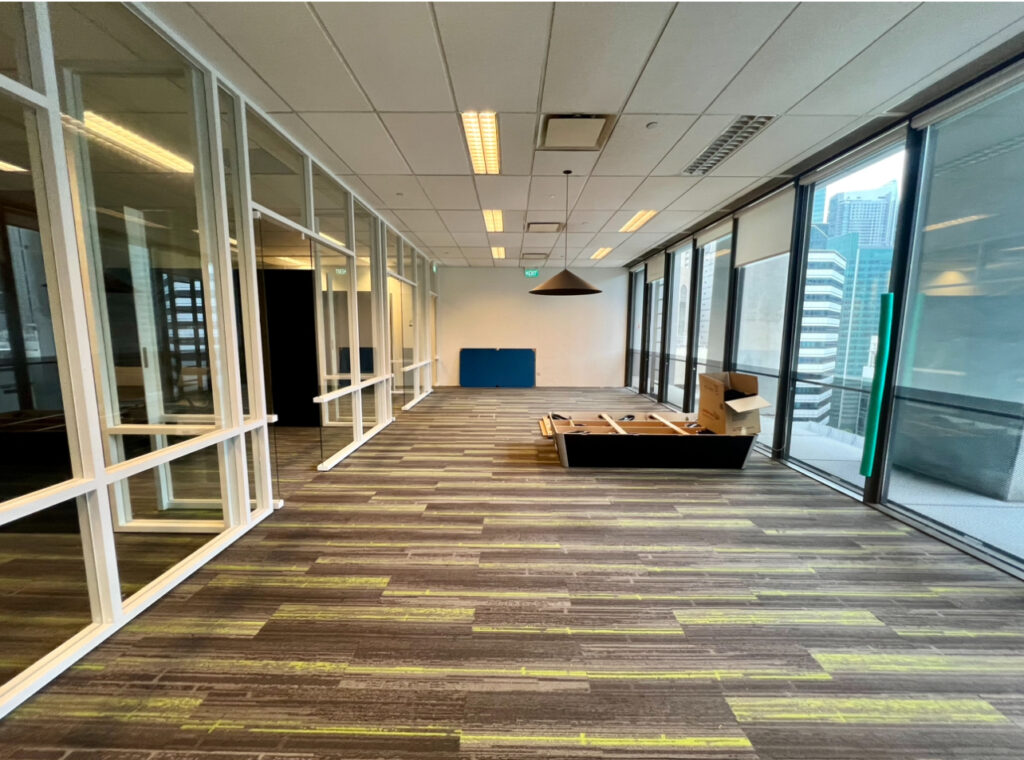 Empty modern office space with glass walls, ceiling lights, a boxed furniture set, and large windows showing city buildings outside.