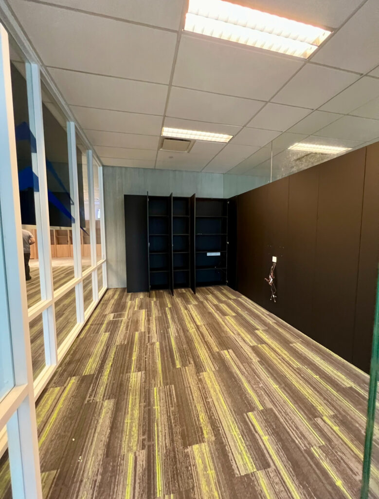 Empty office room with striped carpet, black shelving units against a dark wall, glass partition on the left, and exposed cables on the right wall.
