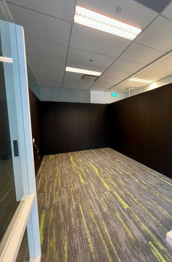 Empty office room with dark walls, grey carpet with green streaks, glass door partially open, and ceiling lights on.