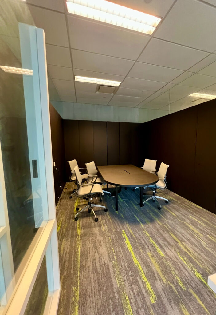 A meeting room featuring a dark oval table surrounded by four white office chairs on a grey carpet with green accents, viewed through an open doorway.