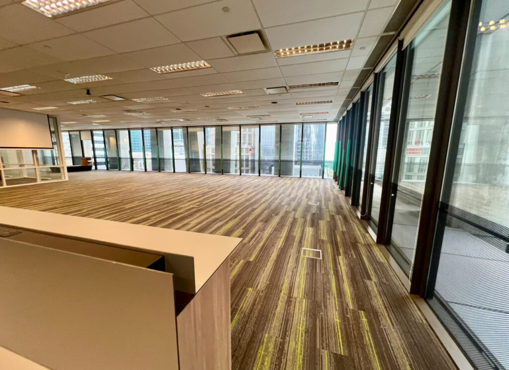 Modern empty office space with carpeted floor, large windows, and overhead lighting. Some office furniture visible in the foreground.