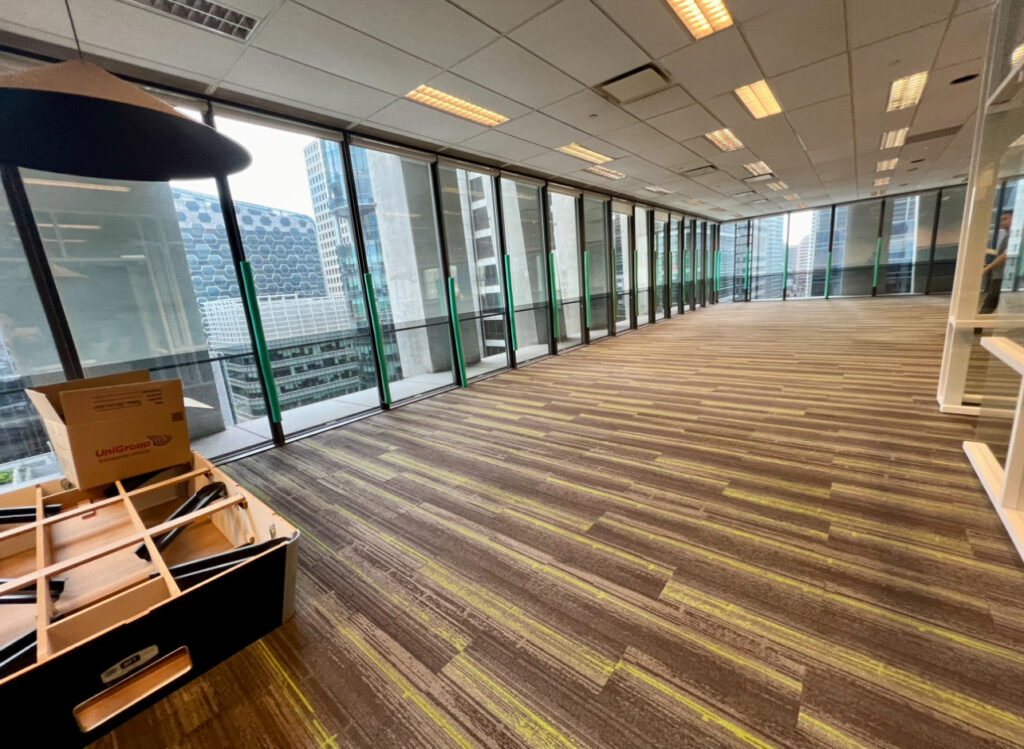 An empty office space with striped carpet flooring, floor-to-ceiling windows overlooking city buildings, and a partial view of a desk or table being assembled in the foreground.