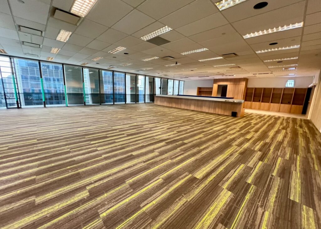 Spacious empty office with wood-patterned flooring, ceiling lights, floor-to-ceiling windows, and a wooden reception desk on the right-hand side.