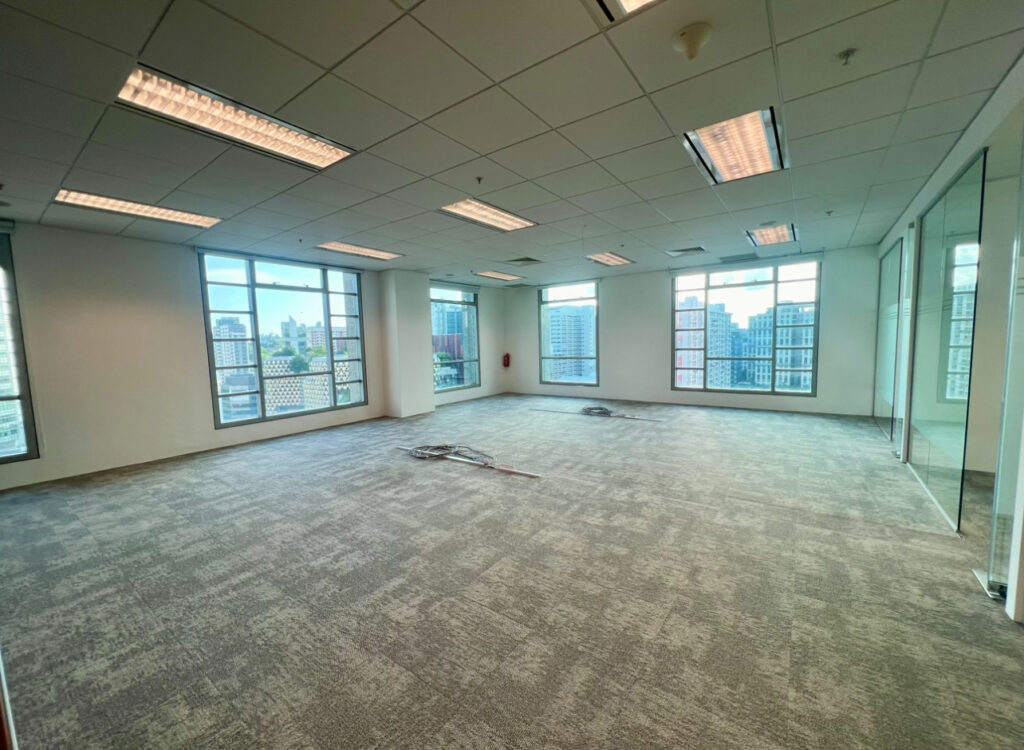 Empty office space with carpeted floor, large windows, overhead fluorescent lights, and some cables lying on the ground. City buildings are visible through the windows.