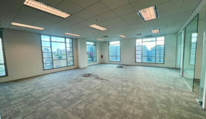 Empty office space with carpeted floor, large windows, overhead fluorescent lights, and some cables lying on the ground. City buildings are visible through the windows.