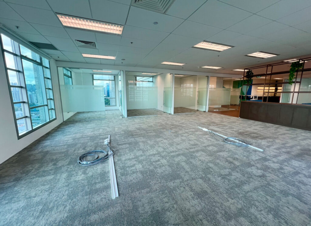 Spacious, empty office with carpeted floor, large windows, glass-walled rooms, and exposed wiring on the floor; city buildings visible outside.