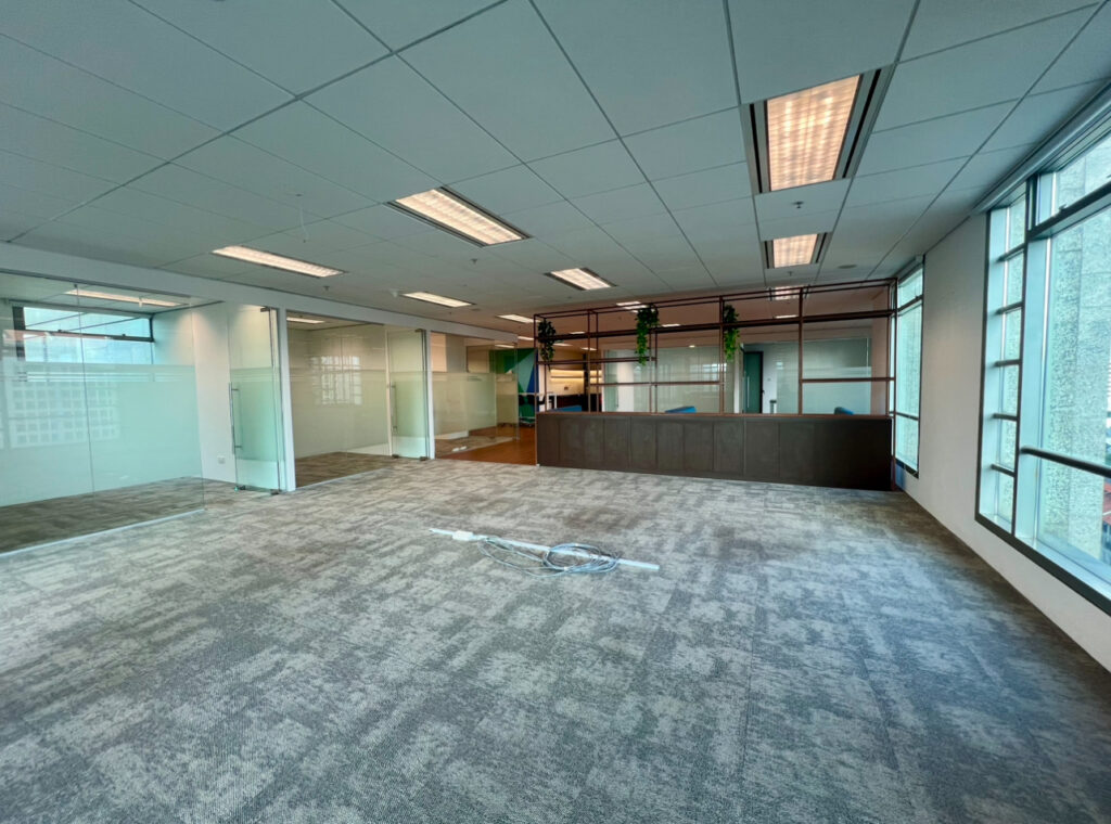 Empty office space with carpeted floor, overhead fluorescent lighting, glass-walled rooms, and a dark wood reception desk; loose wires are visible on the floor.