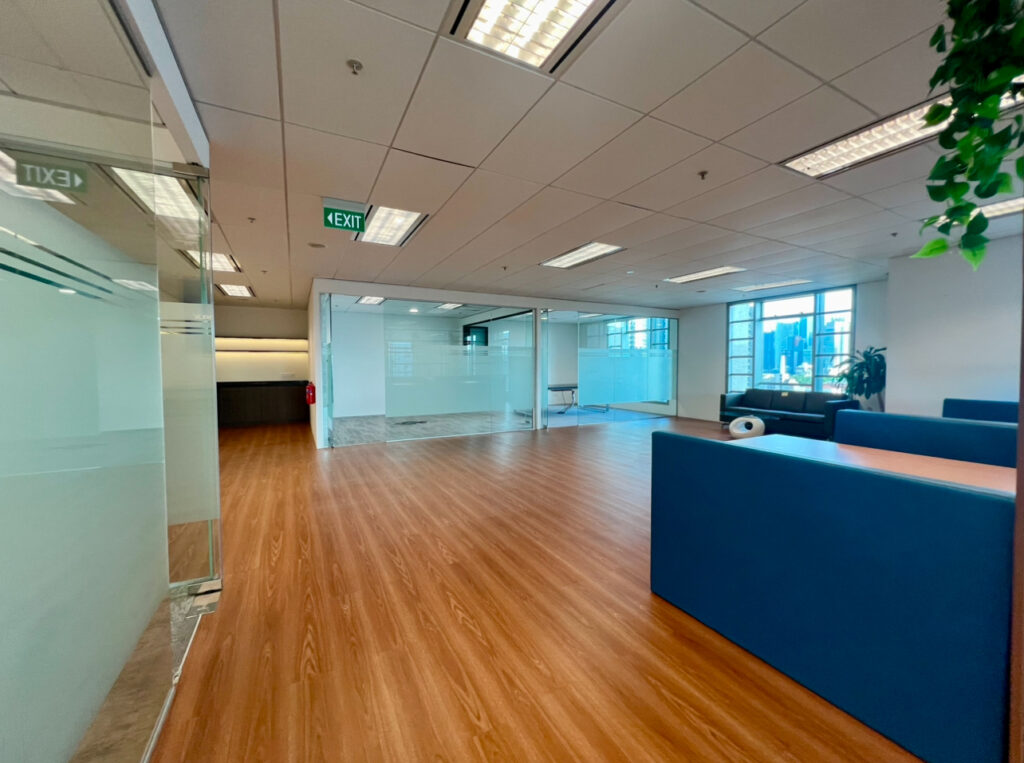 Modern open-plan office space with wooden flooring, blue seating, glass-walled meeting rooms, ceiling lights, exit signs, and a few plants near large windows.