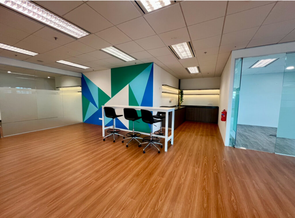 Modern office space with wood flooring, a high table with four chairs, geometric feature wall, glass partitions, and overhead lighting.