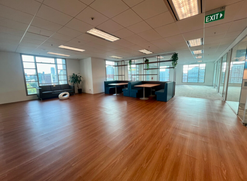 Spacious office area with wooden flooring, blue booth seating, a black sofa, large windows, plants, and glass-enclosed rooms; an exit sign hangs from the ceiling.