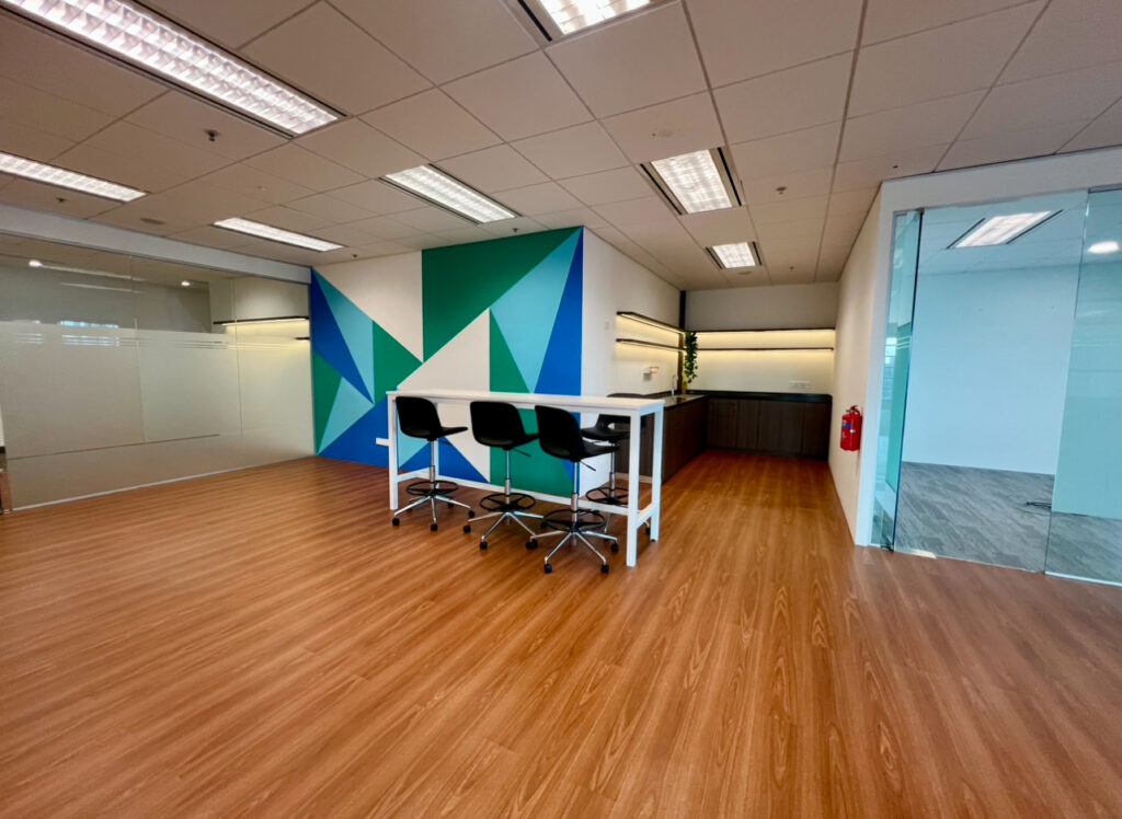 Modern office space with wood flooring, a high table with four chairs, geometric wall art, and glass-walled rooms.