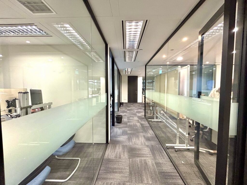 Modern office corridor with glass-walled meeting rooms on both sides, grey carpeted floor, and overhead fluorescent lighting.