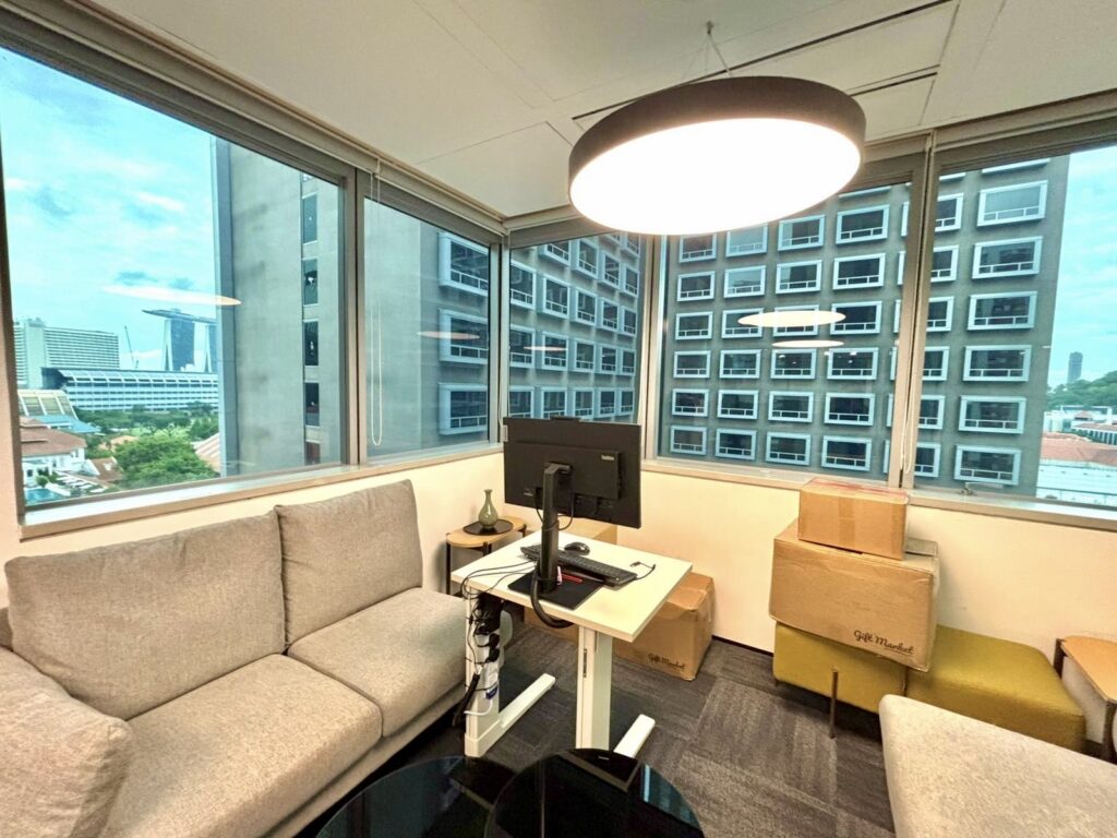 Modern office with a standing desk, computer monitor, grey sofa, unpacked boxes, and large windows overlooking city buildings.
