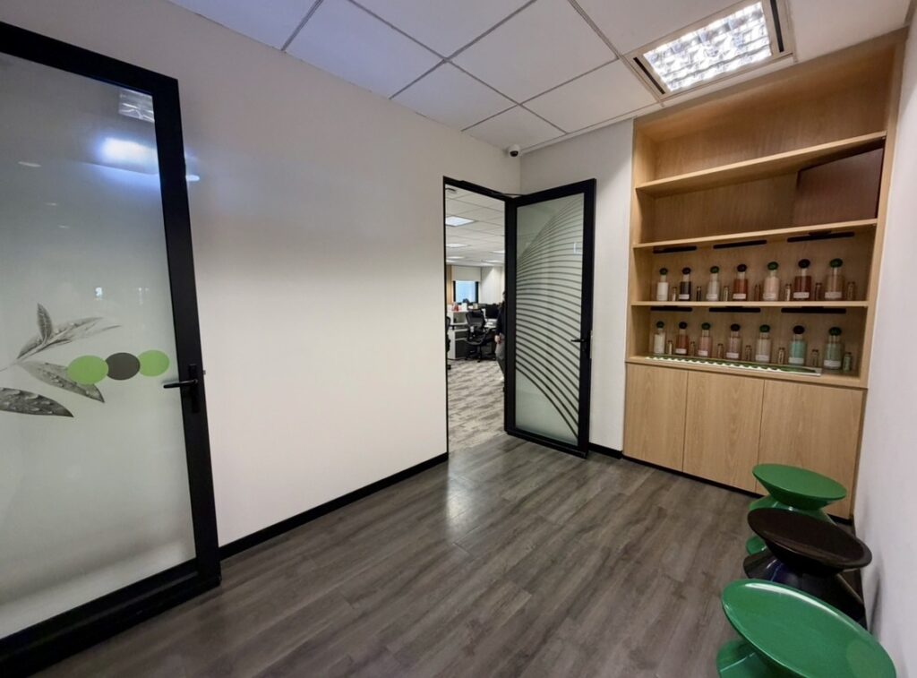 Hallway with wooden flooring, a shelf holding bottles, green stools, and a glass door leading to an office area with desks and people.