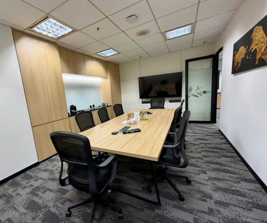 Modern conference room with a long wooden table, eight chairs, a wall-mounted TV, and wooden cupboards. There are drinks and notepads on the table.