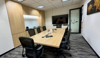 Modern conference room with a long wooden table, eight chairs, a wall-mounted TV, and wooden cupboards. There are drinks and notepads on the table.