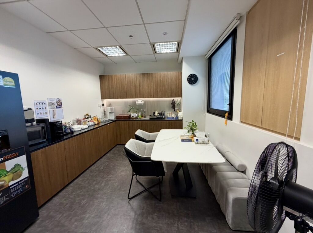 Office kitchen with a marble table, chairs, a bench, kitchenette with appliances, plants, a wall clock, and a standing fan. Large window on the right, ceiling lights above.