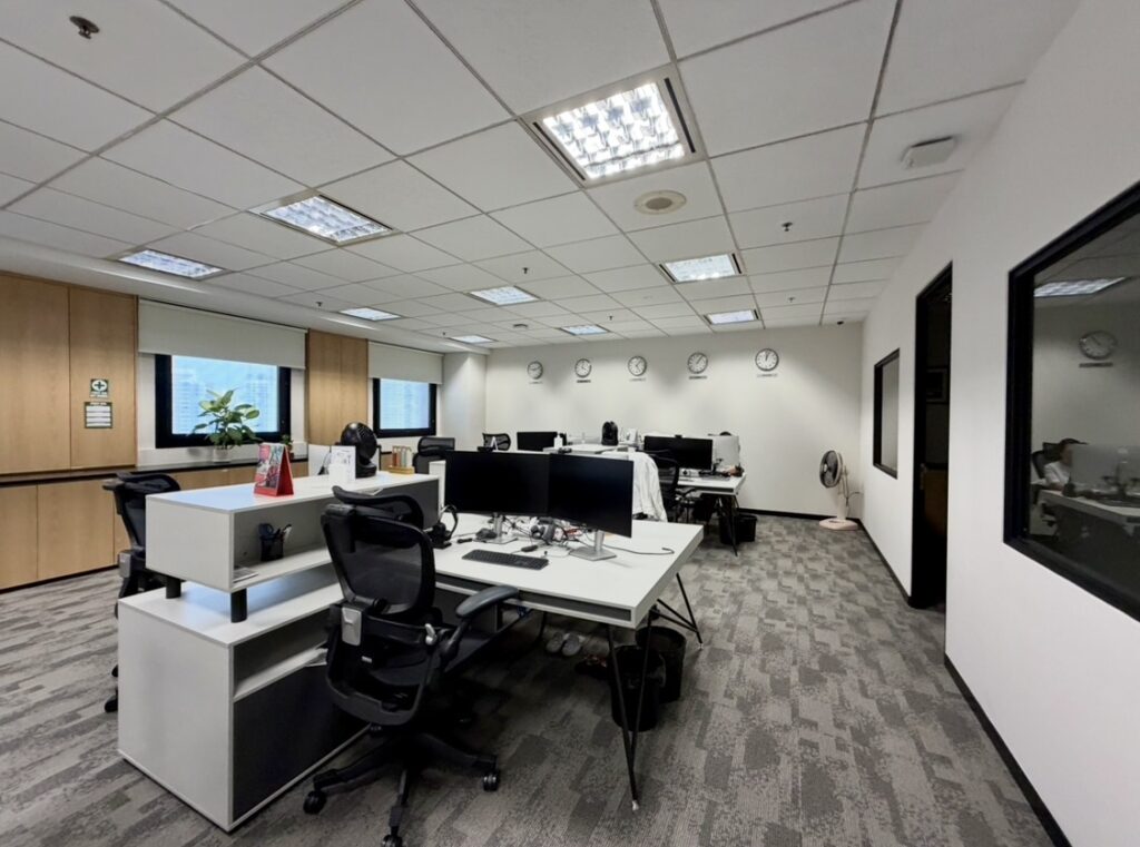 Modern office with empty desks, chairs, computers, and clocks on the back wall, bright fluorescent lighting, and neutral-coloured decor.