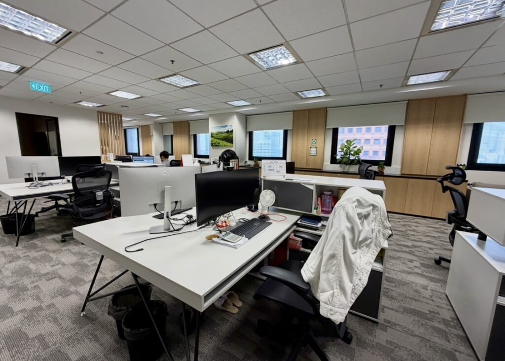 Modern office with several workstations, computers, chairs, and personal items. Large windows provide natural light. A white jacket hangs on a chair in the foreground.