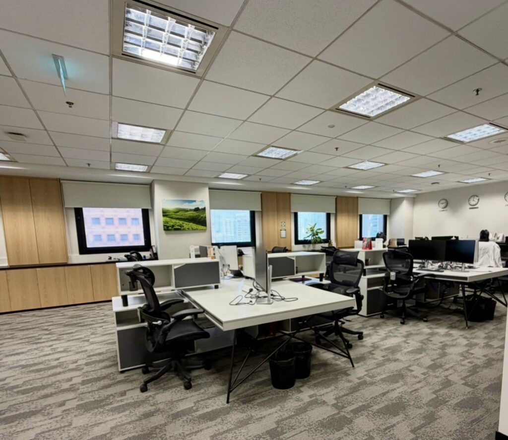 Modern open-plan office with several empty desks, chairs, and computer monitors, neutral carpeting, wood panelling, and large windows along one wall.