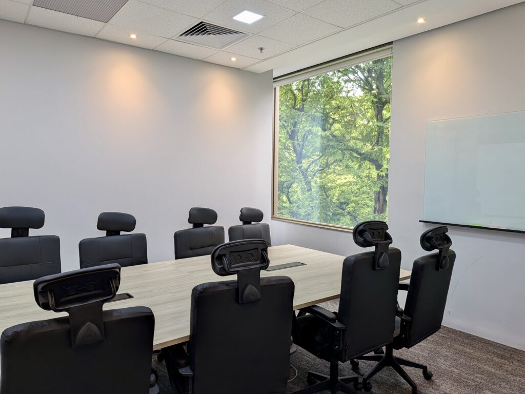 Modern conference room with a long table, black office chairs, large window showing greenery, and a whiteboard on the wall.