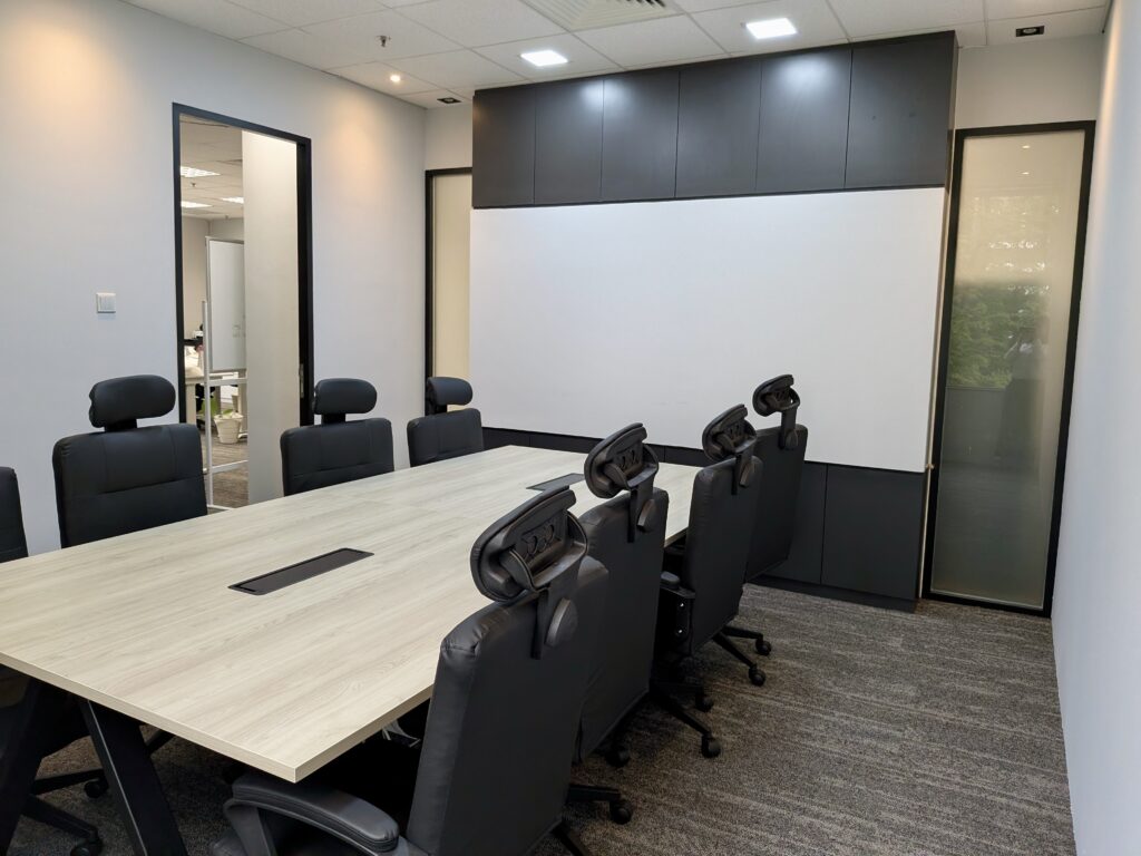 A modern conference room with a rectangular table, eight black chairs, a blank whiteboard, and neutral-coloured walls and carpet.